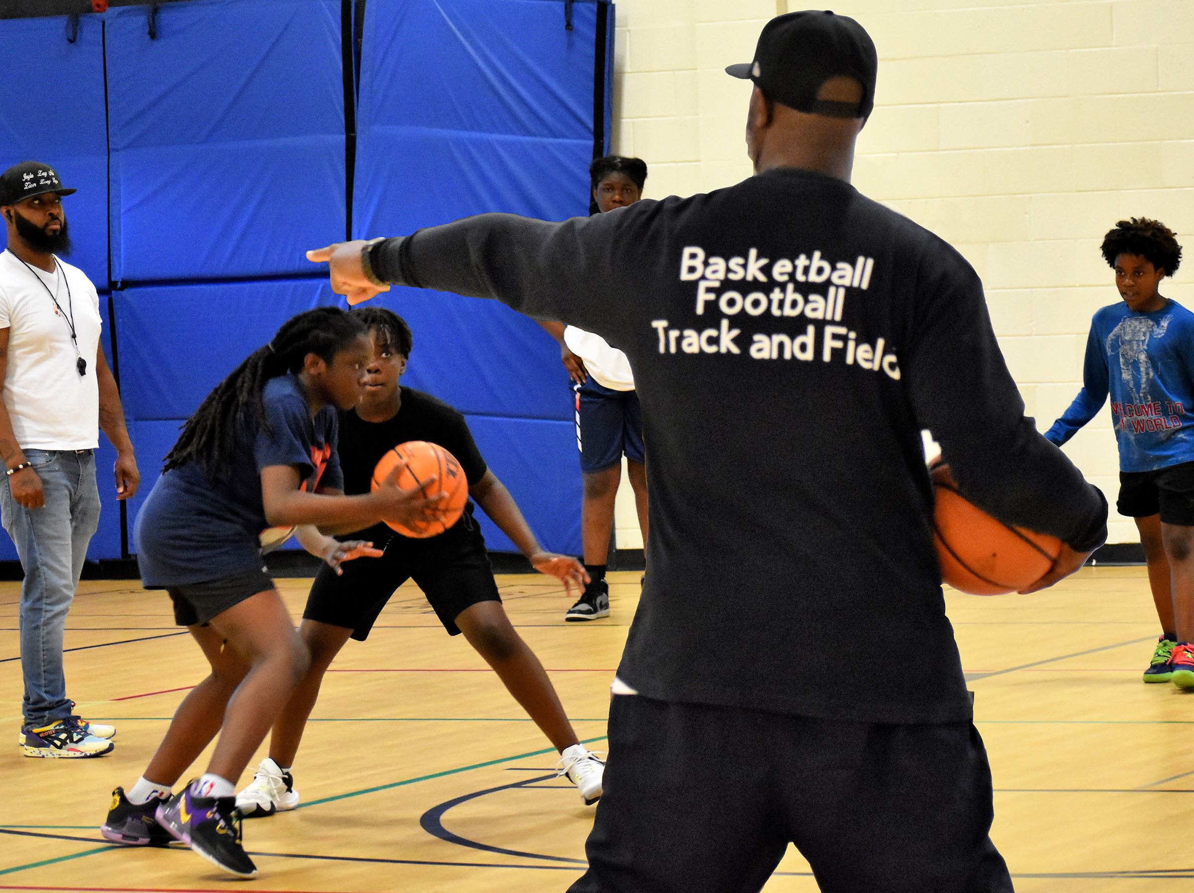 A coach in a black t-shirt stands on a gym floor pointing at adolescent boys playing basketball