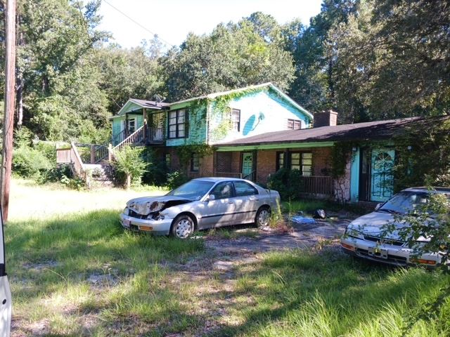 An abandoned house overgrown with weeds a vines. An abandoned car sits in front of it.
