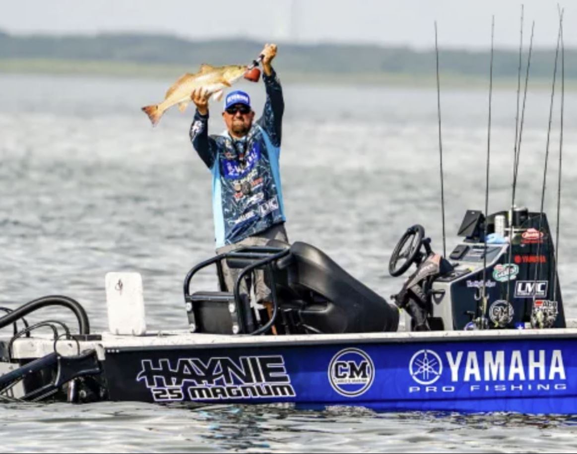 A man in a boat in the middle of the water holds a large redfish above his head, showing it off.