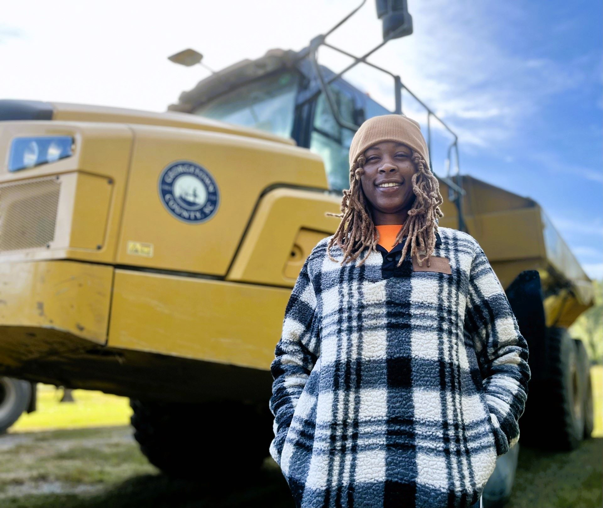 A young black woman in a knit cap stands in front of a large, yellow off-road articulated dump truck