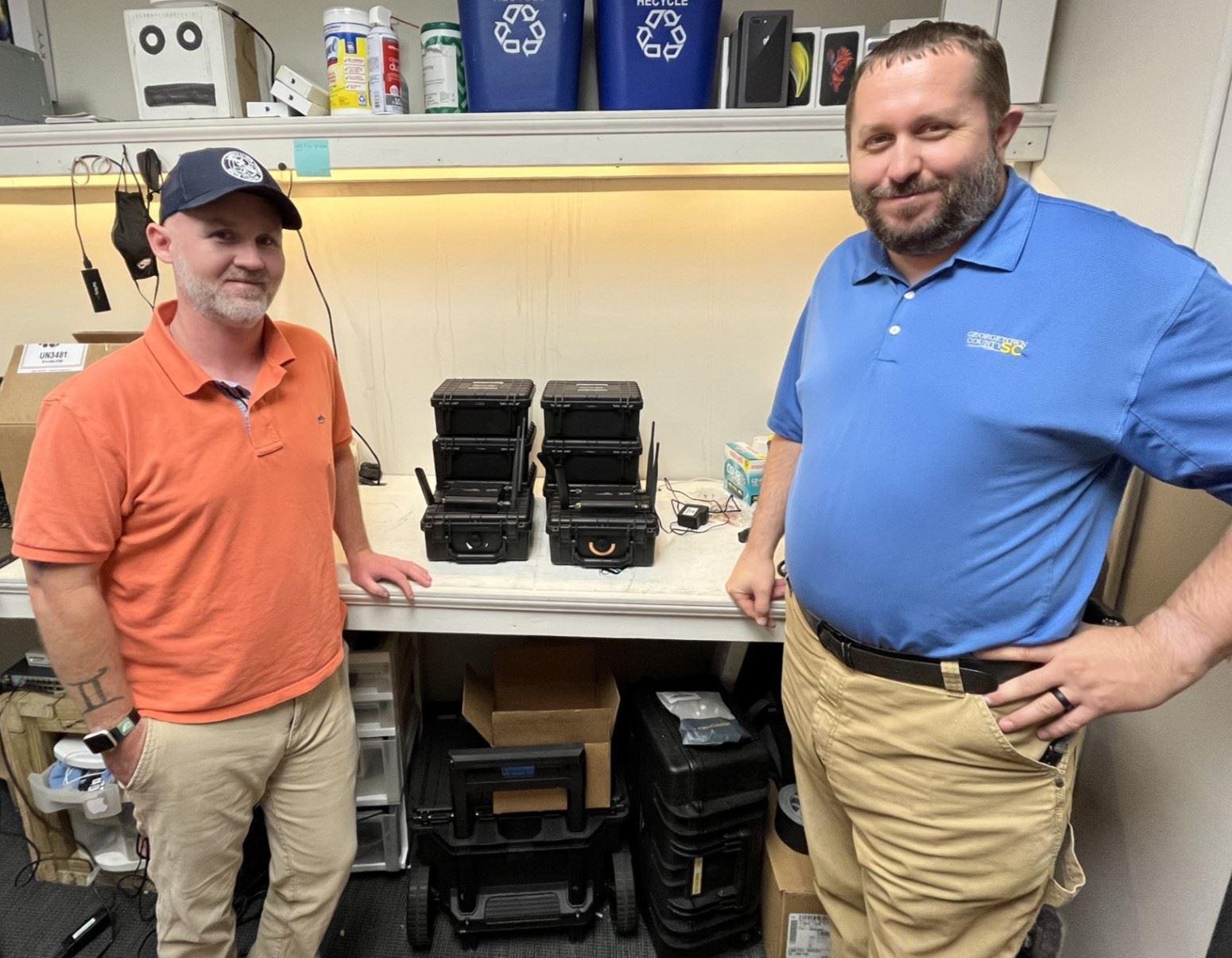 Two male MIS staff members stand on either side of a stack of cradlepoint routers
