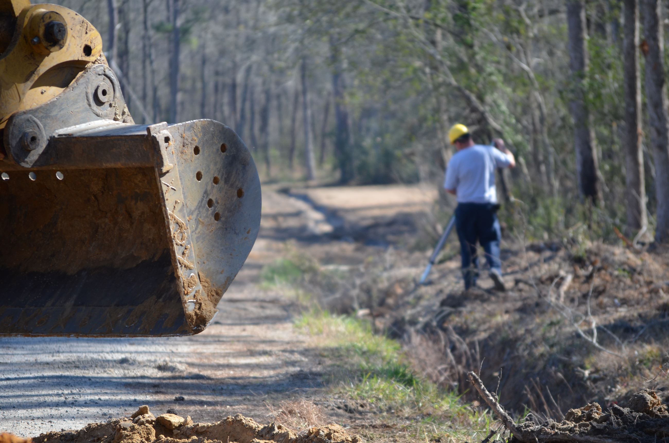 A man in a hardhat works in a ditch with a backhoe visible in the foreground