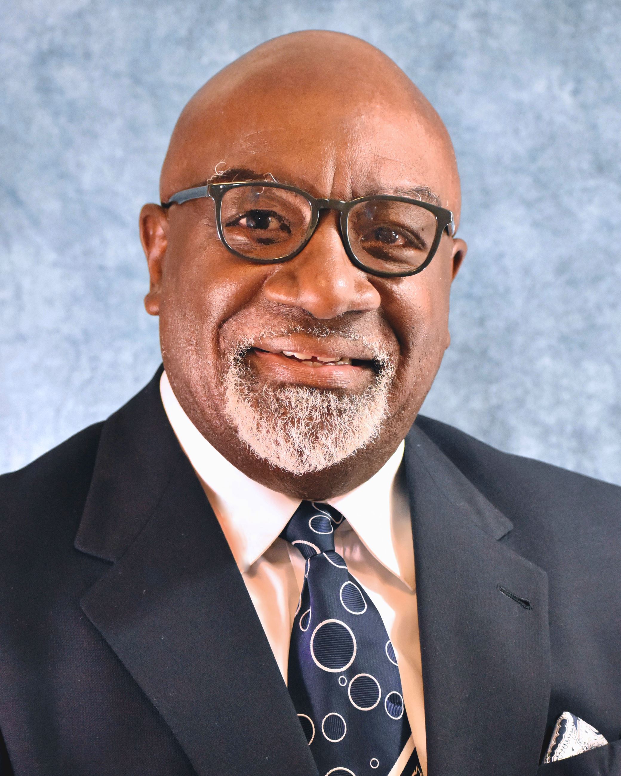 Ernie Cooper, a bald African American man with a white goatee wearing glasses and a suit, smiling.