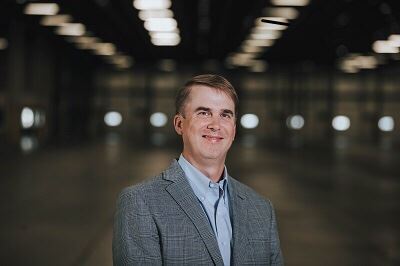 A man wearing a blue shirt and grey blazer, smiling.
