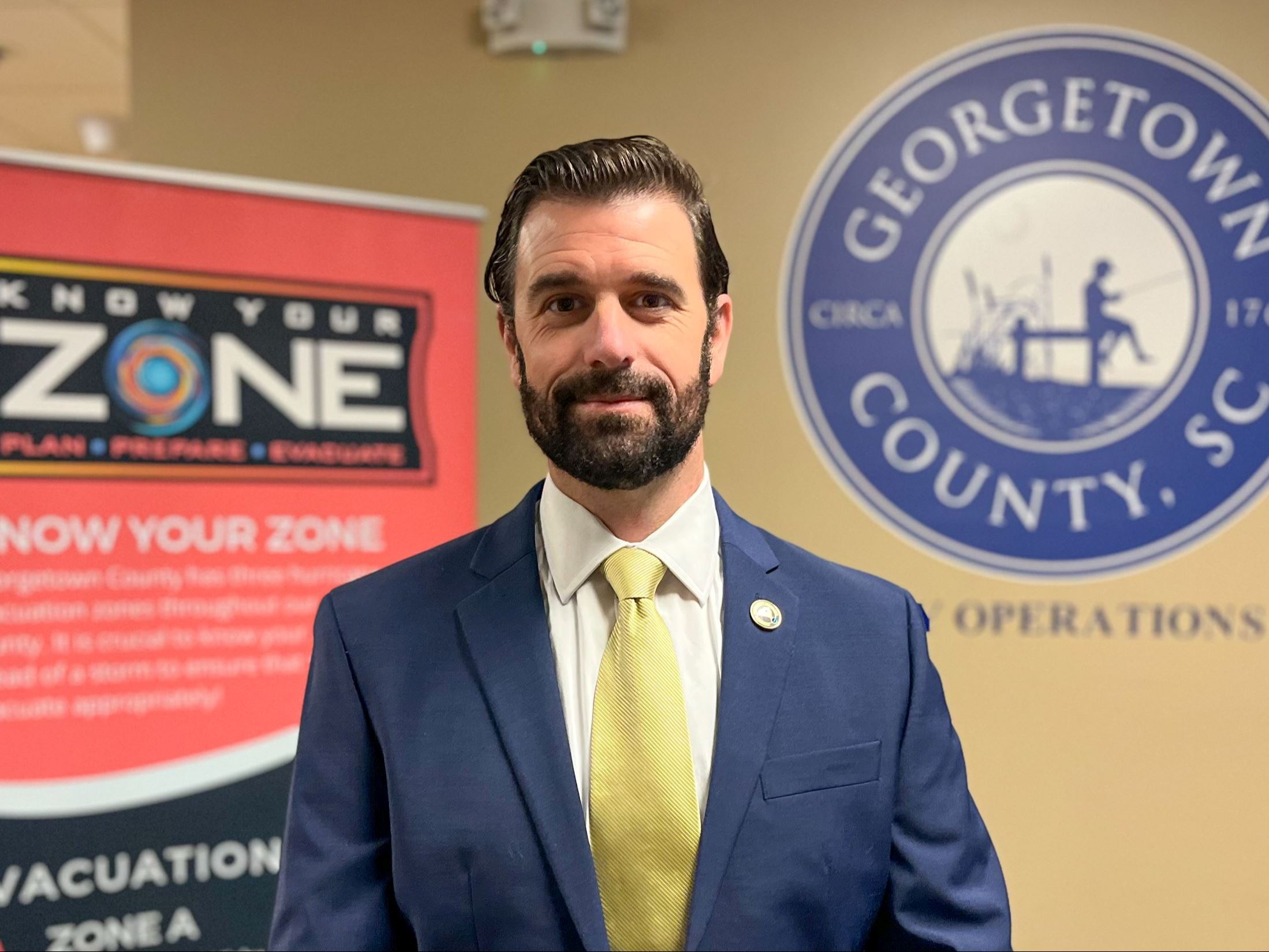 A bearded man in a suit stands in front of a wall emblazoned with the Georgetown County logo