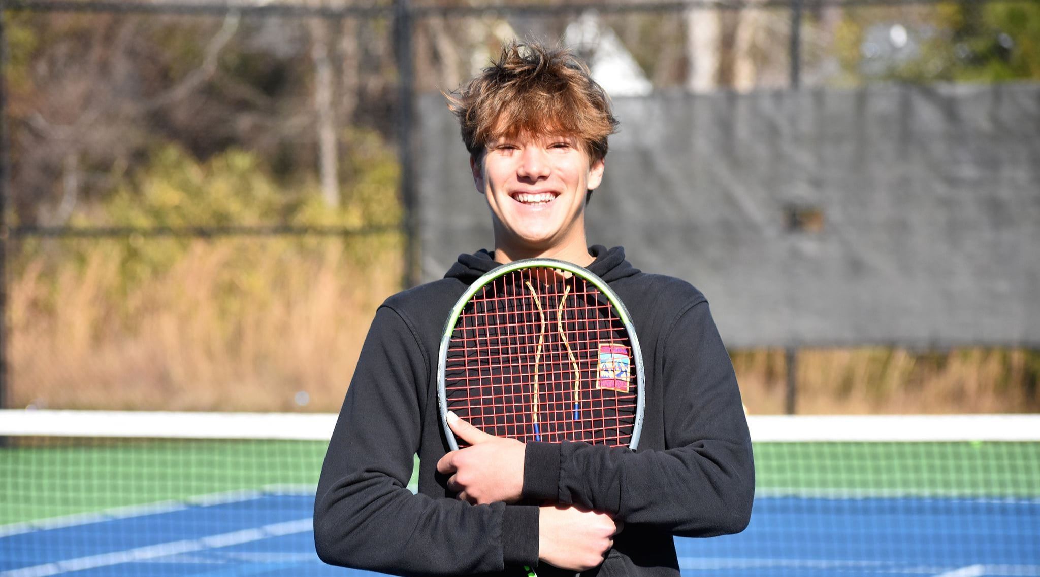 A teenage boy in a black hoodie holds a tennis racquet against his chest