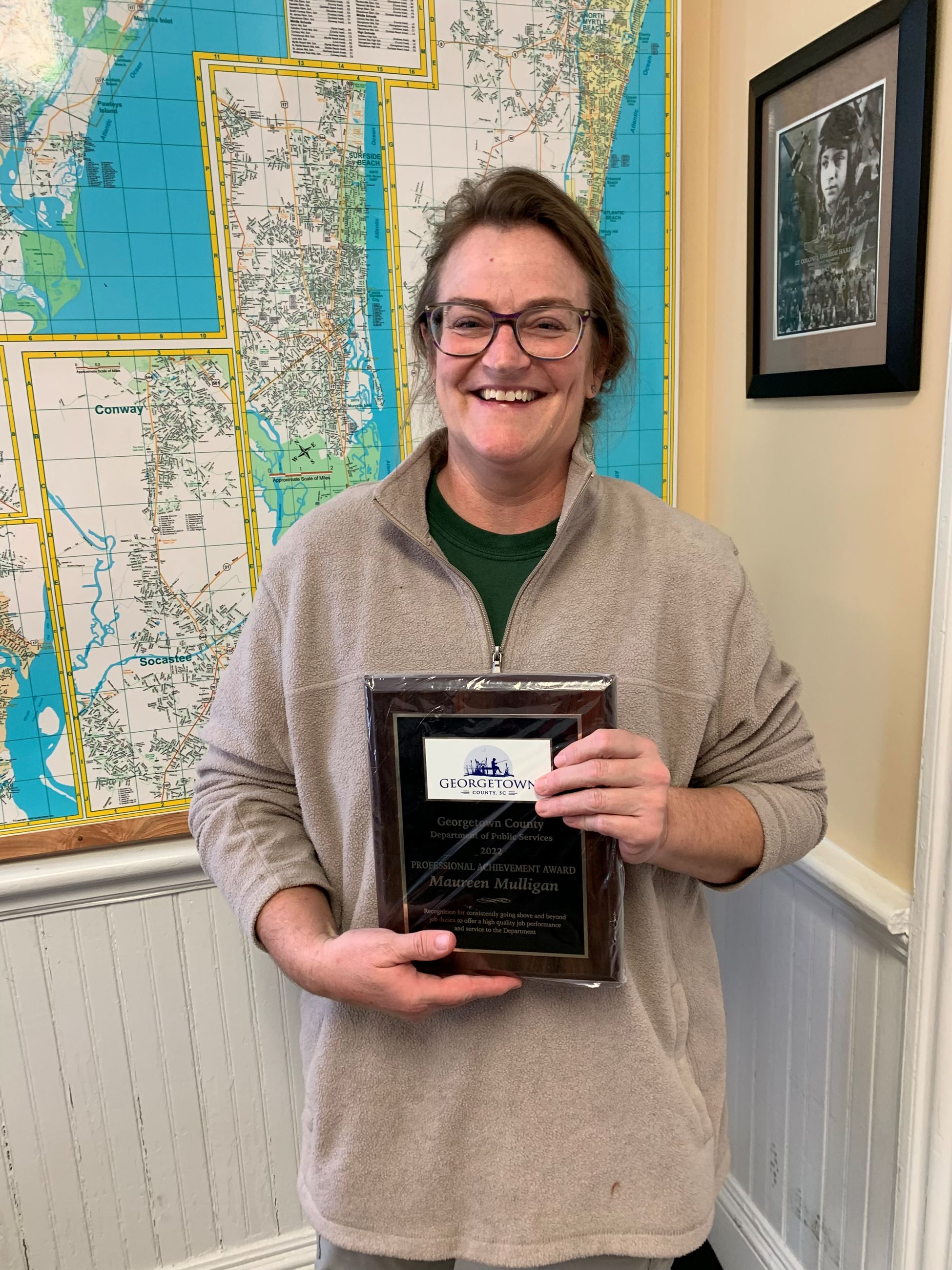Woman wearing glasses and light brown zip-up pullover, holding an award plaque and smiling.